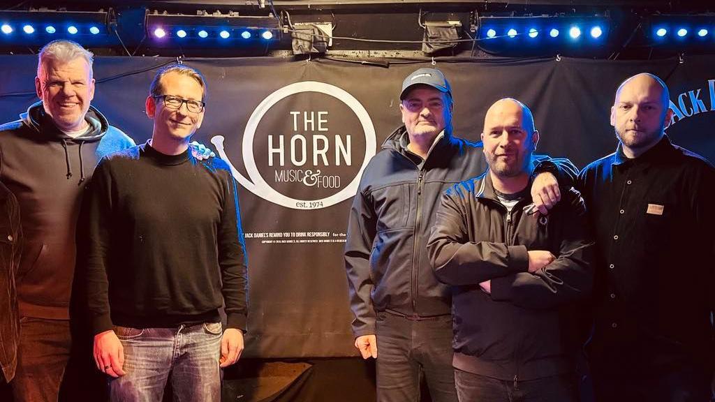 Five men stood together, dressed in dark clothing. Behind them is a sign for The Horn pub.