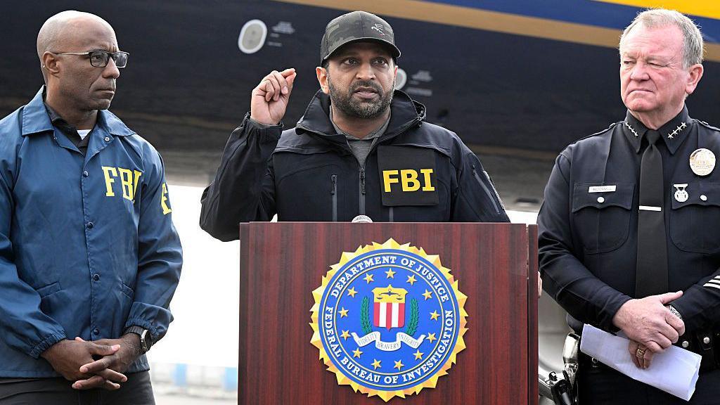 Kash Patel wearing an FBI jacket speaking at a podium with an FBI agent standing to his right and a police officer to his left.