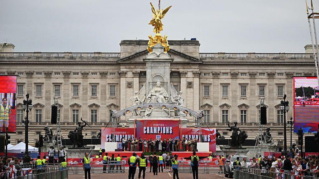 A bandstand is shown in front of Buckingham Palace with signage that reads European Champions.