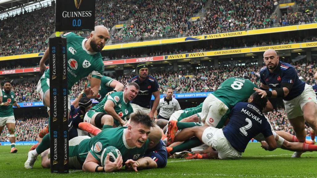 Dan Sheehan of Ireland dives over the try line with a ball in his hand to score his team's first try whilst under pressure from Maxime Lucu of France (who is obscured from view) during the 2025 Six Nations Championship match between Ireland and France at the Aviva Stadium in Dublin in March. An Irish teammate of Sheehan is behind him and other players of both sides look on with the stadium view in the background