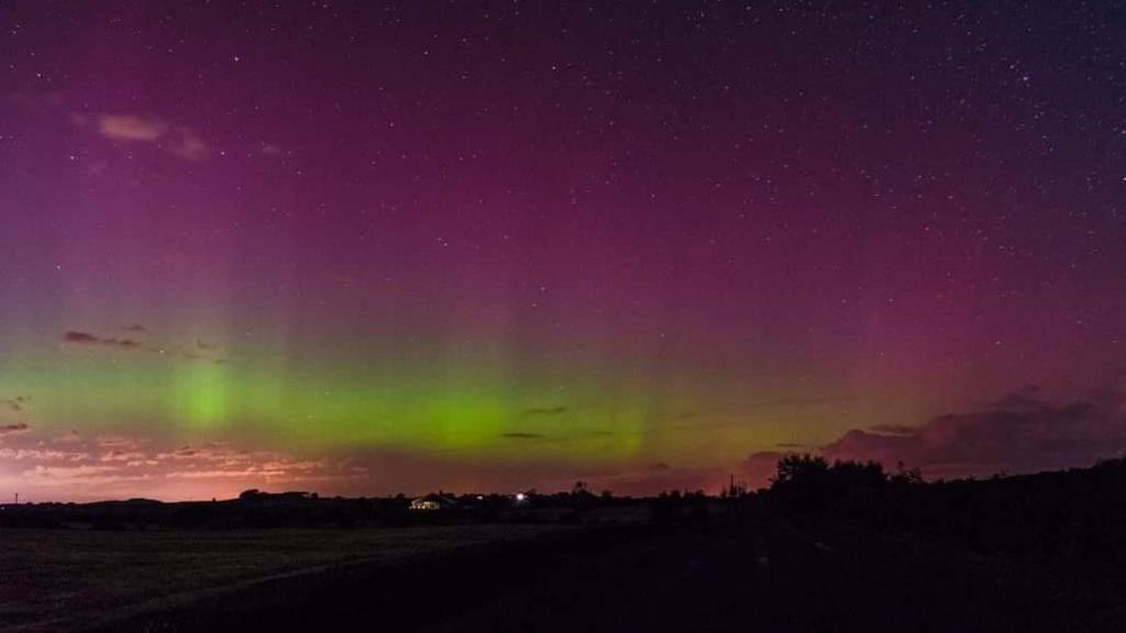 Sky with stars Northern Lights in purple and green over some trees and a building with lights in the distance