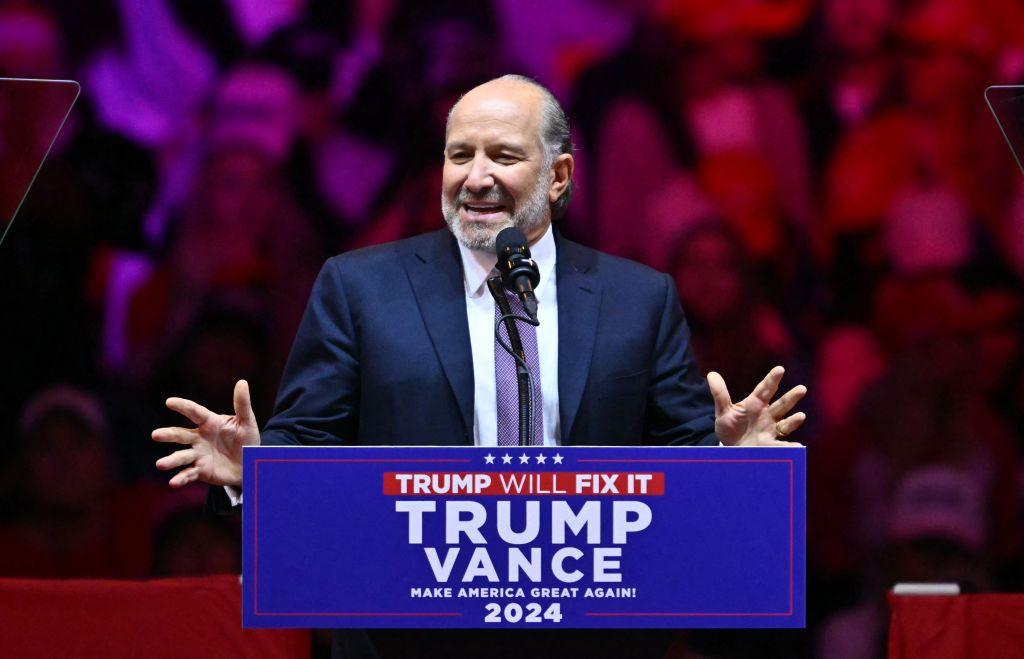 Howard Lutnick speaks while gesturing with both hands in front a lectern at at a Trump rally in Madison Square Gardens in October. He has grey hair and a beard, and wears a navy suit, white shirt and purple checkered tie.