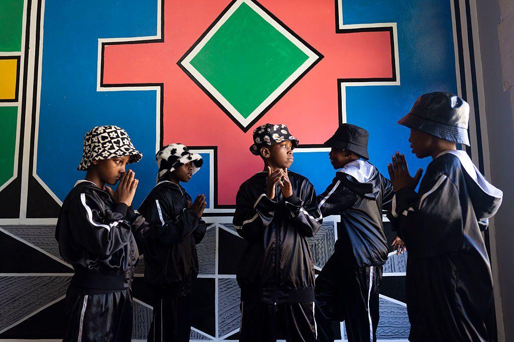 Children in dark tracksuits and monochrome bucket hats stand together in room in front of a mural.