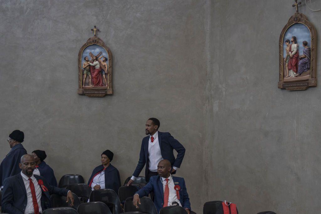 Men dressed in matching dark suits and red tie wait in seats. Above them are depictions of the stations of the cross.