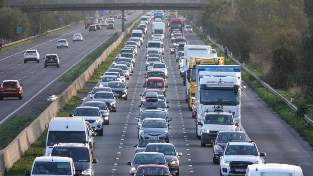 Vehicles queue in a traffic jam as rain on the M5 motorway near Locking. The traffic on the right hand carriageway driving towards the camera is very busy, the other side is not. There is a bridge over the road in the background.