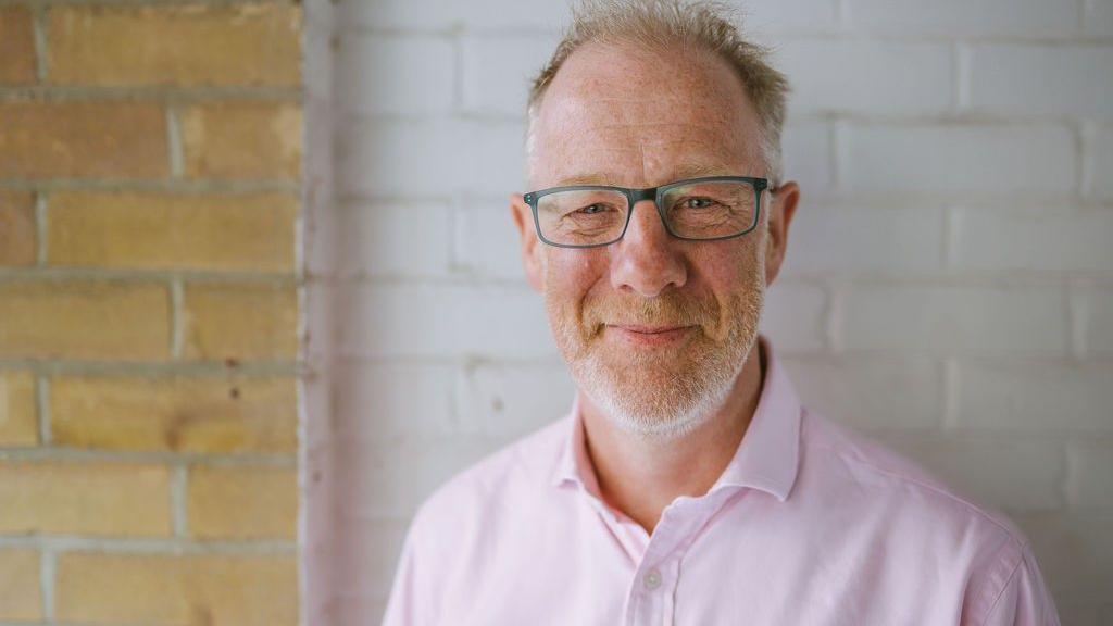 Ben wearing a pastel pink shirt with ginger and grey stubble wearing blue rimmed glasses smiling at the camera. He is standing in front of a brick wall. Some of it has been painted white.