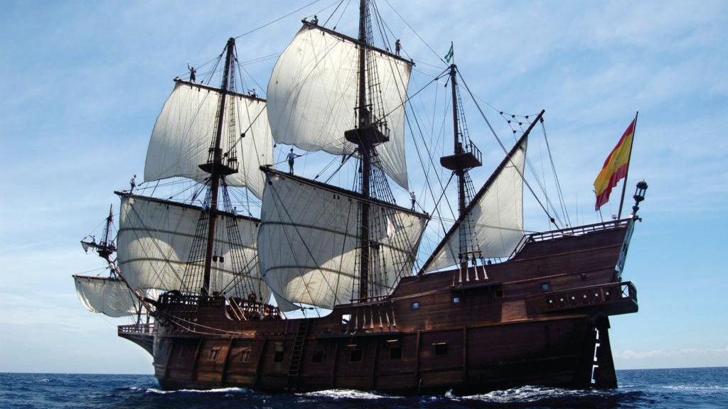 A huge 17th century galleon ship with a wooden hull and decks, six visible white sails and flying a Spanish flag whilst launched on the sea.