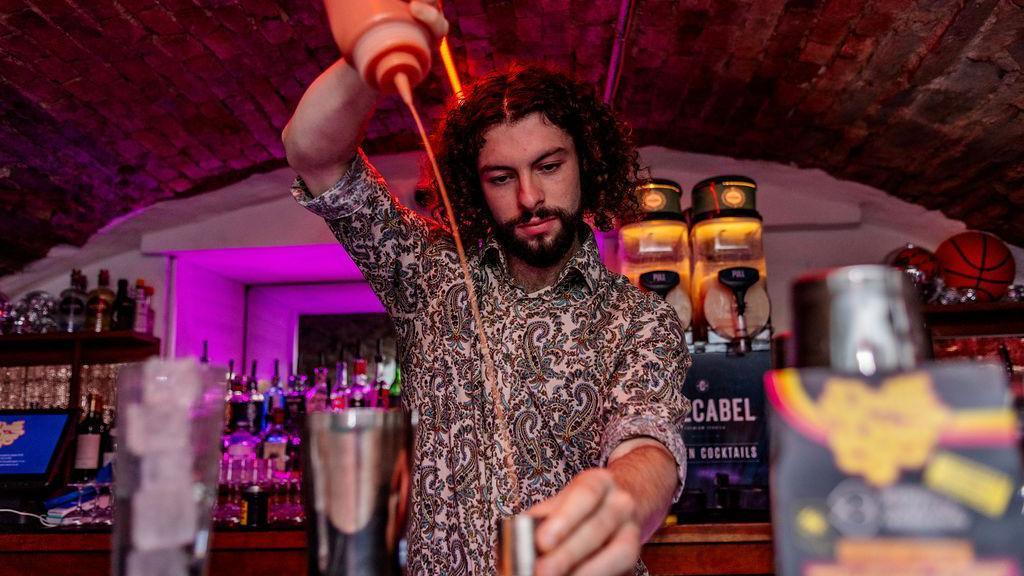 A man in a paisly-patterned shirt pours liquid into a cocktail measuring glass in a bar with multiple coloured bottles behind him. He was photographed during the closing event for Bristol Cocktail Week which took place at Death Disco