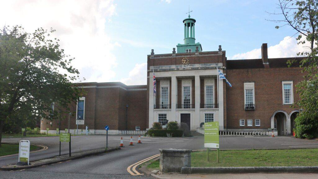The main front of Hertford County Hall, with a green pillared table on of a wide 1930s style brown brick building. It has tall long windows at the front, a gold clock and two flags. In the foreground is a green sign for Hertfordshire County Council