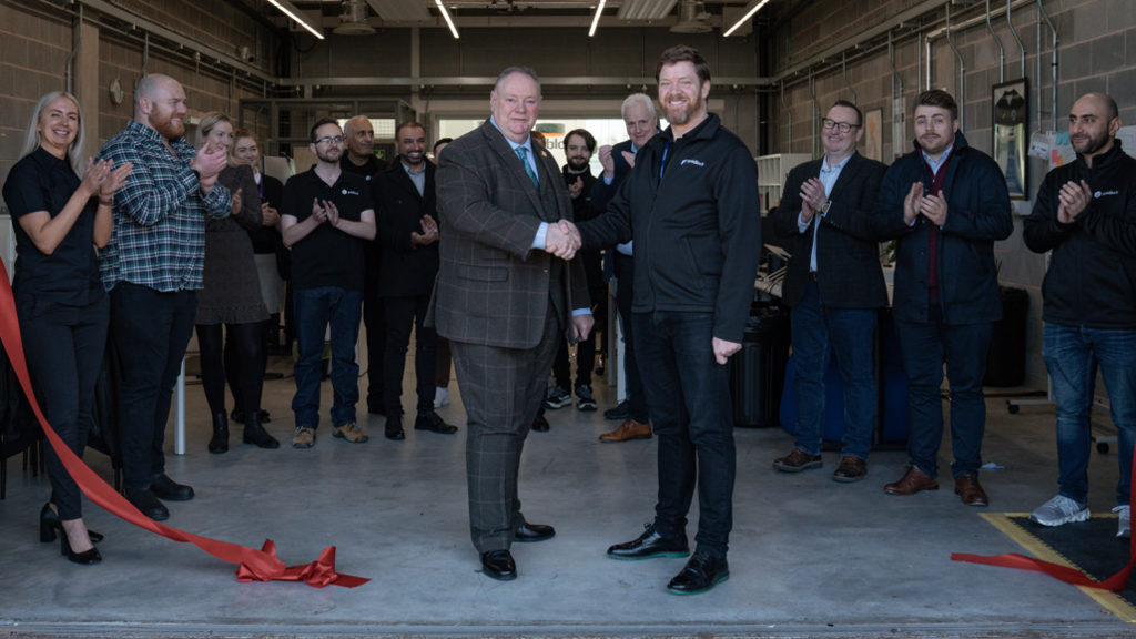 Council leader Stephen Simkins and Stephen Kines shake hands after a red ribbon is cut at the expanded premises. Behind them is a crowd of about a dozen men and women clapping.