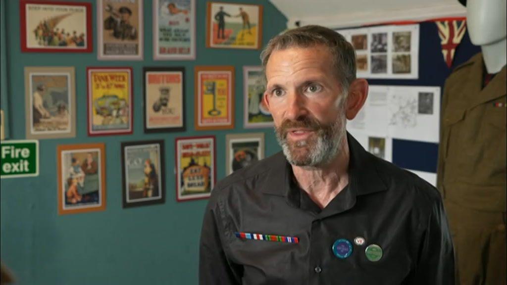 A man with a black shirt and badges looks into the camera. He has short hair and a beard and behind him on a wall there is a series of adverts and slogans from during World War Two.