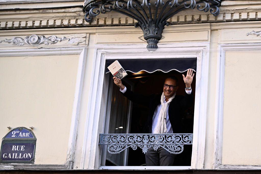 Laureate of the Prix Goncourt literary prize, Algerian writer Kamel Daoud waves from a window of Drouant Restaurant Paris, holding up his book. A street sign for Rue Gaillon can be seen on the wall to the left of the window - Monday 4 November 2024

