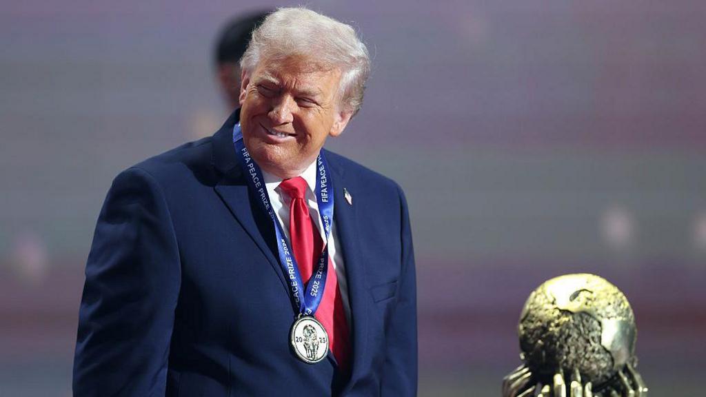 Donald Trump looks to his right and smiles as he wears a smart blue jacket, pink tie, and has a Fifa Peace Prize medal on a ribbon around his neck