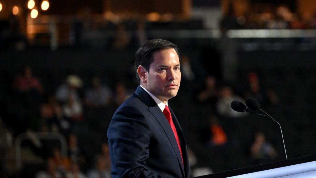 Marco Rubio stands at a podium while wearing a red tie and blue jacket. the background is dark and he is seen at an angle