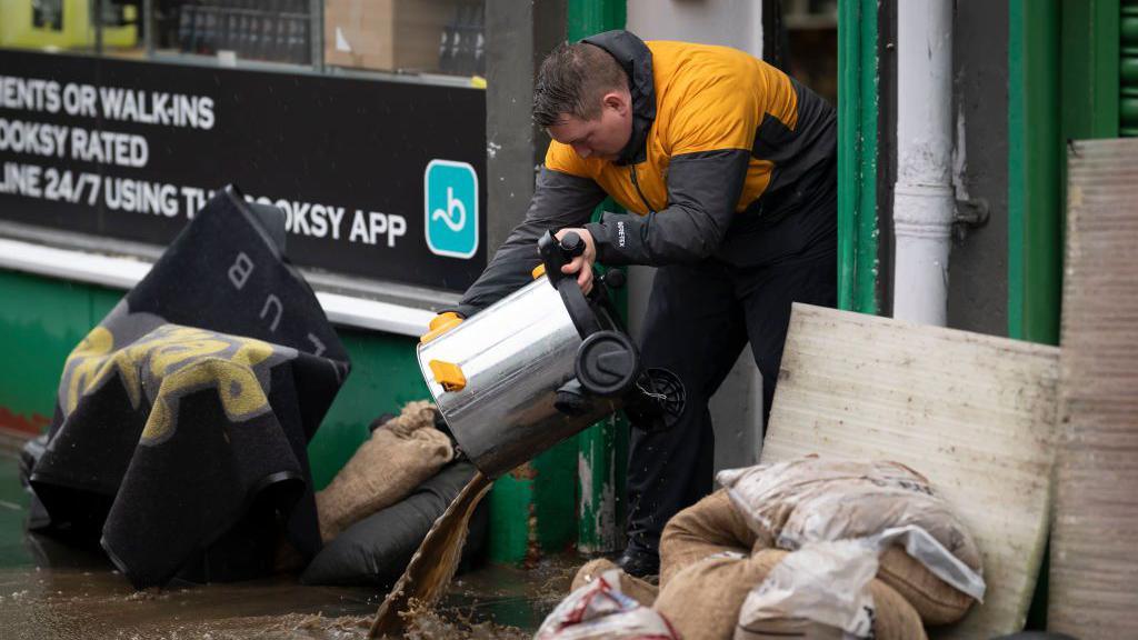 A man bails water out of a business in Pontypridd. There is rainwater in the street, amongst sandbags.