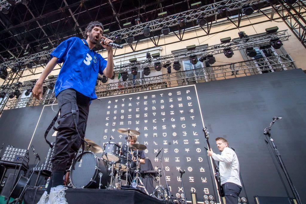 Sergio Pizzorno in a blue shirt and black trousers, Ian Matthews sat behind a drum kit and Chris Edwards in a white shirt playing guitar,  of Kasabian perform at Lucca Summer Festival on July 06, 2022 in Lucca, Italy.