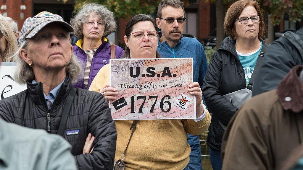 A woman in a crowd of people holds a sign saying "USA: Throwing off tyrants since 1976"
