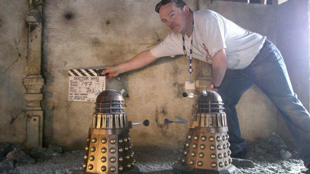 Mike Tucker holds a clapper board that says "Doctor Who" close to two model Daleks, as they prepare to film an effects shot for Day Of The Doctor.