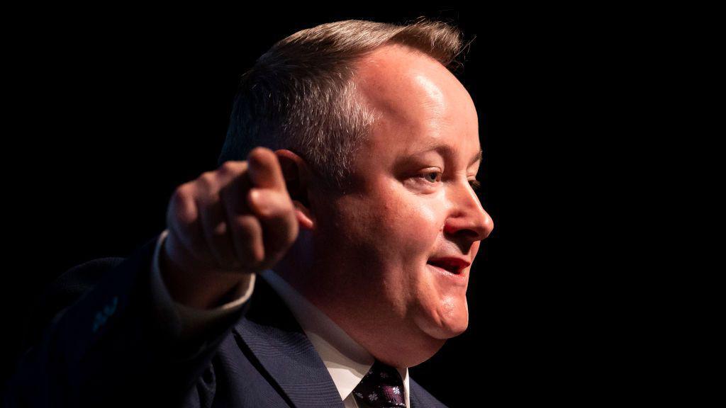 Darren Millar is pointing towards the camera while looking away at a right angle towards an audience. He is wearing a suit, tie and shirt, and is stood before a dark background.