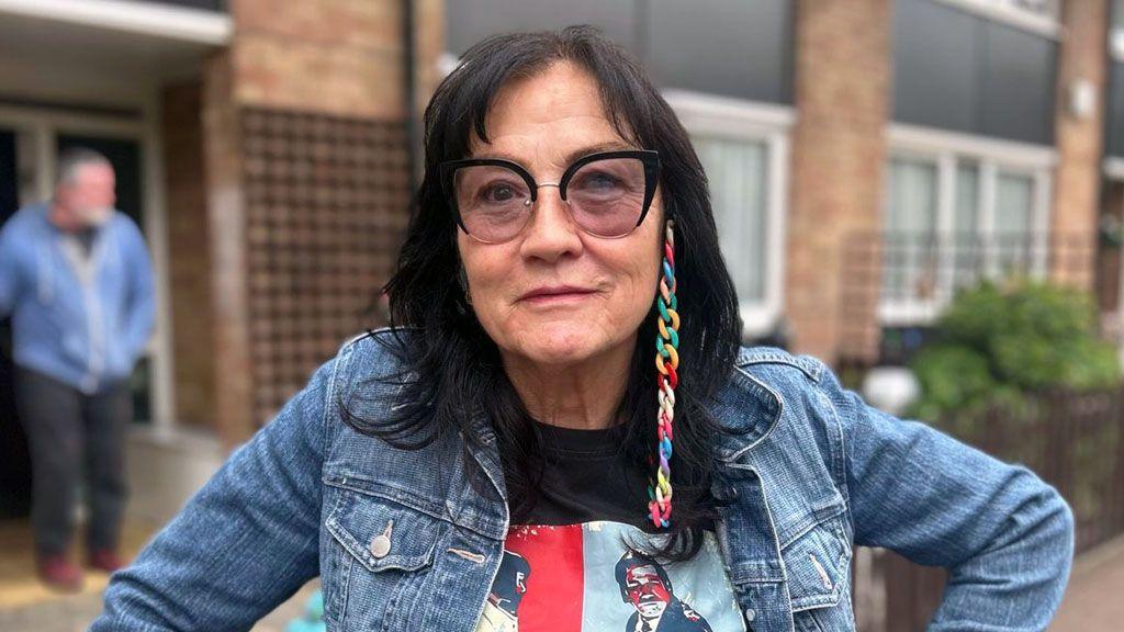Headshot of Tracey. She has long dark hair with a fringe, and large black-framed glasses. She is wearing a black t-shirt with a print of the characters from Pulp Fiction, and a blue denim jacket. She is pictured standing in front of a row of houses.