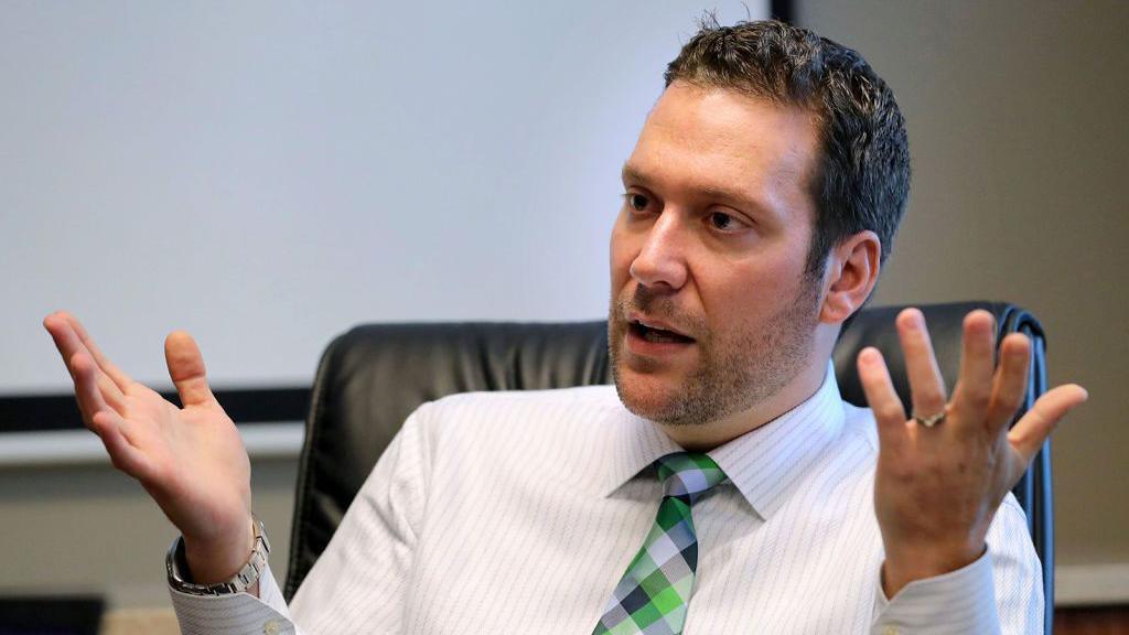 Joel Greenberg, who has short dark brown hair and facial hair, and is wearing a white shirt with light black stripes and a chequered tie, gestures with both his hands during a 2019 interview