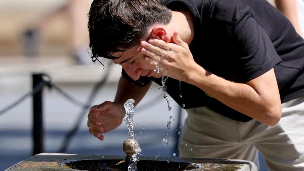 A man leans over a concrete water fountain, splashing water onto his face