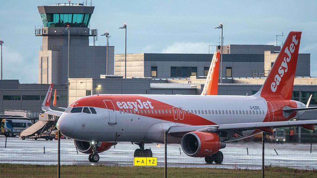 Awyren oren a gwyn EasyJet o flaen adeiladau maes awyr Bryste a ddiwrnod heulog, er bod y tarmac yn wlyb.