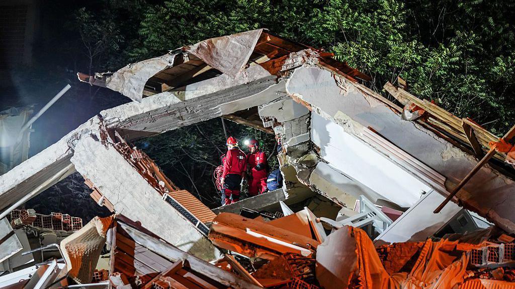 Emergency workers can be seen through a square frame that is being created by a massive chunk of collapsed building.