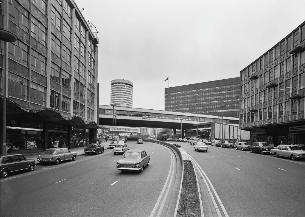 A black and white image of Birmingham's Ringway Centre in the 1970s, with cars seen driving down the wide route. The city's rotunda tower can be seen in the background with cars parked on either side of the road.