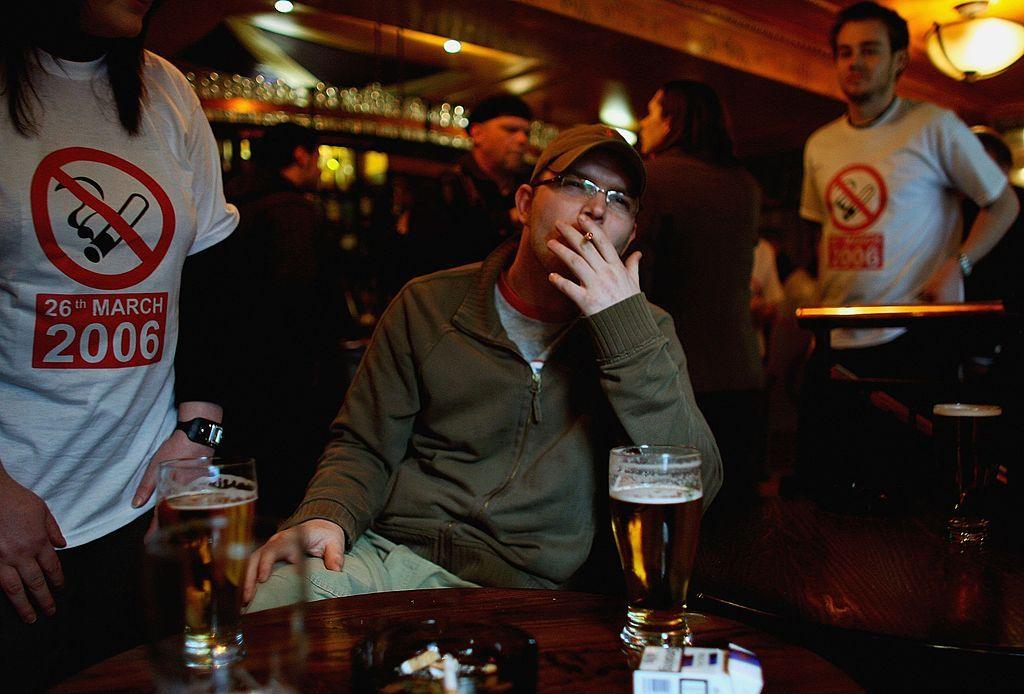 A man wearing glasses, a dark green zipped top a green baseball hat sits smoking in a pub, with pints of beer and a cigarette packet on the table front of him. There are also two people standing wearing white tshirts with a red logo highlighting that the smoking ban will come into effect on 26 March 2006.