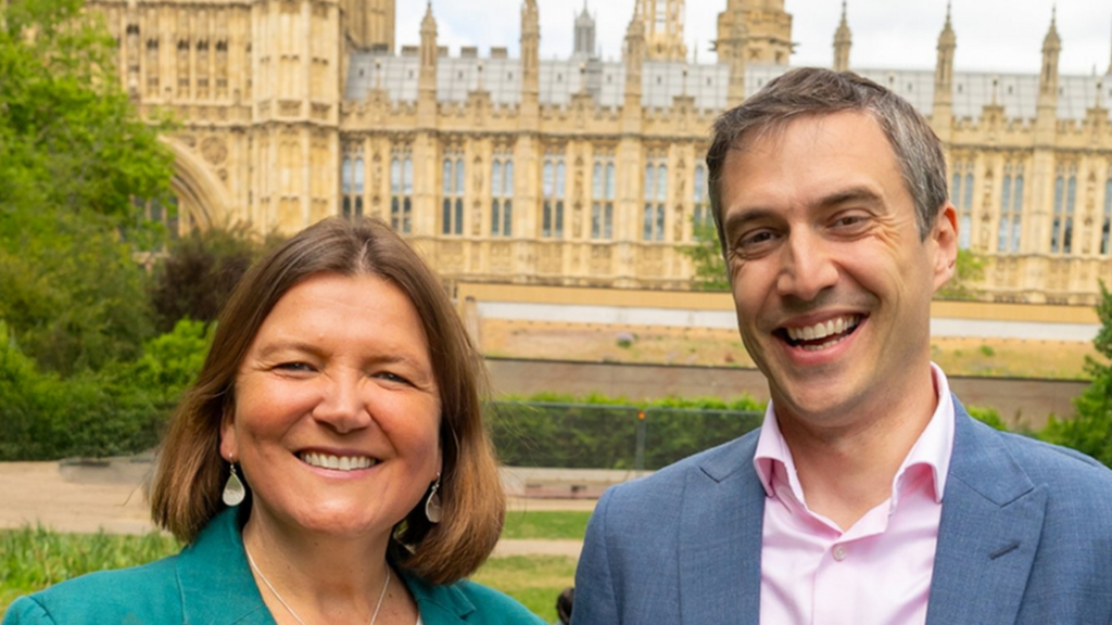 Ellie Chowns and Adrian Ramsay stand outside Parliament. Chowns wears a turquoise jacket with silver jewellery and Ramsay wears a smoke-blue suit with a pink, open-necked shirt.