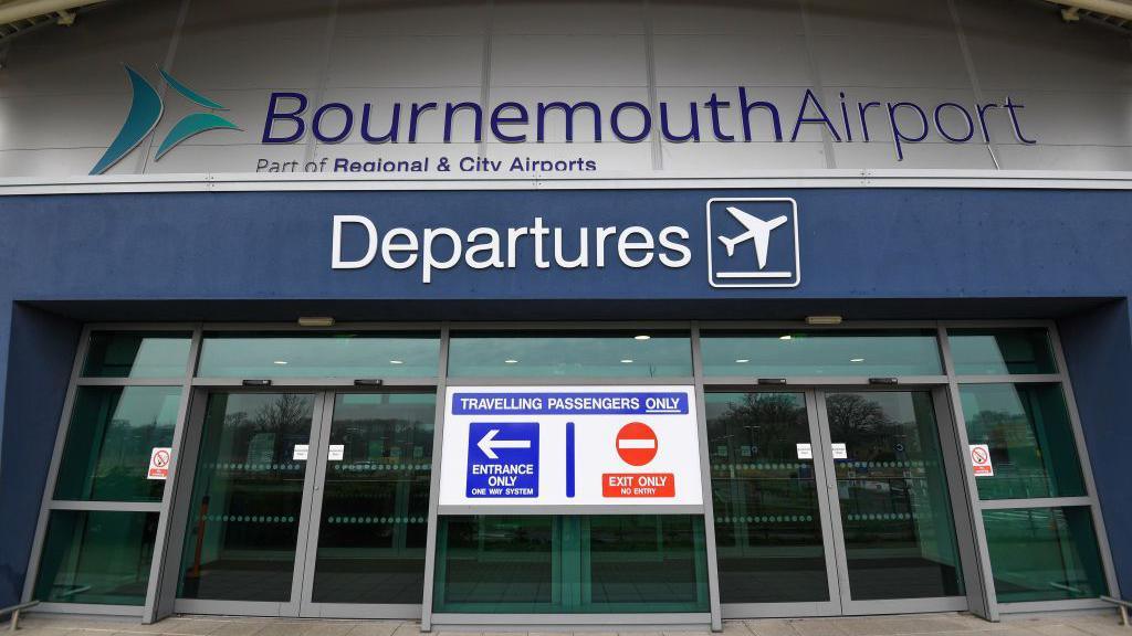 The entrance for departures at Bournemouth Airport - with airport sign in blue above the departures sign in white above glass doors and a sign advising it's for travelling passengers only and which door is the entrance and which the exit.