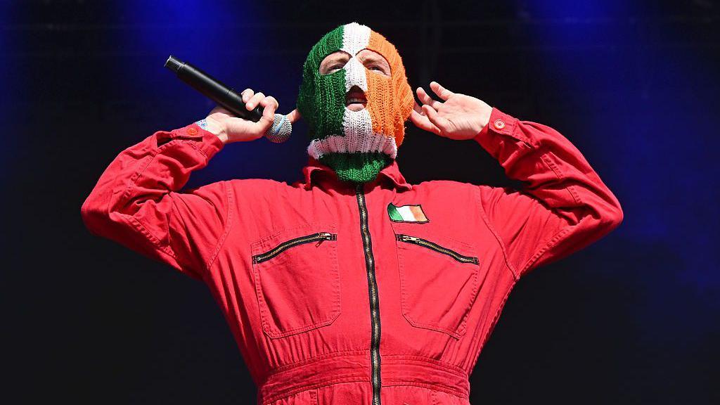 A man wears a red boiler suit with a green white and orange flag on his left chest. He wears a balaclava that is the same colour as the flag. He puts both his hands up to his ears. His right hand holds a black microphone.