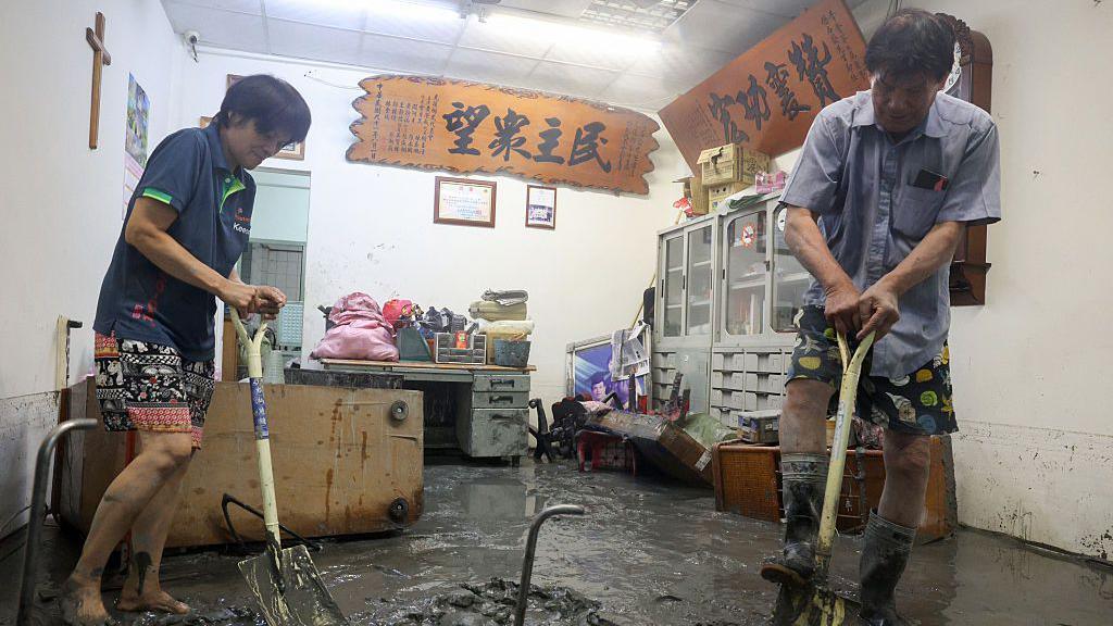 Two residents (a woman on the left, a man on the right) in t-shirts and shorts use spades to clear up the mud within a property in Hualien 