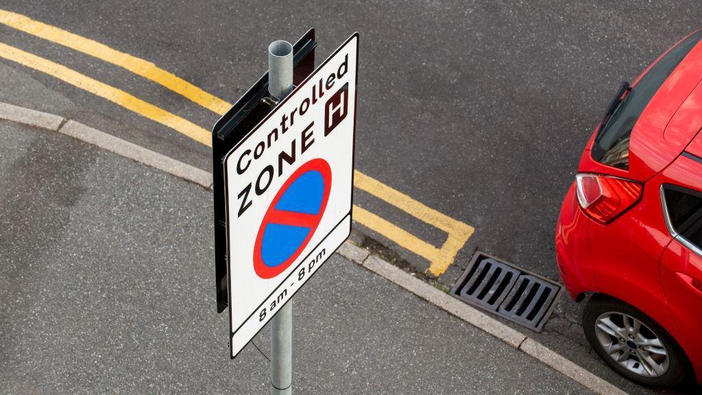 A road sign saying "Controlled zone, 8am-8pm" with a red car stopped next to it.