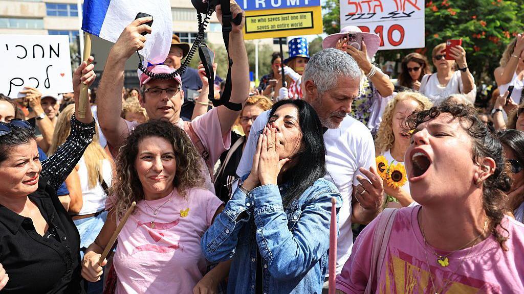 Einav Zangauker, in a blue shirt, holds her hands, palms pressed together, to her mouth. People around her holding signs and taking pictures smile and cheer.