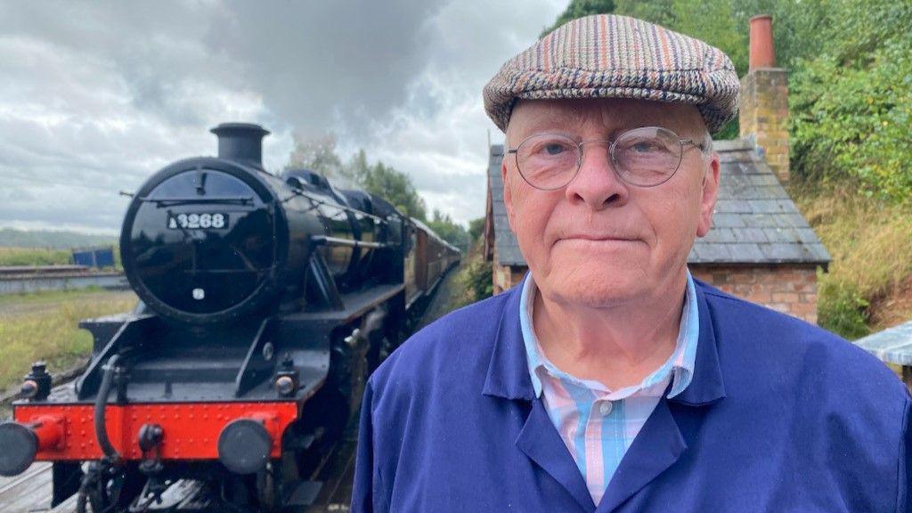 Phil Harris is standing to the right of a black and red steam train. He is wearing a blue jacket with a blue and pink shirt underneath. He has a tweed hat on his head, and is wearing glasses.