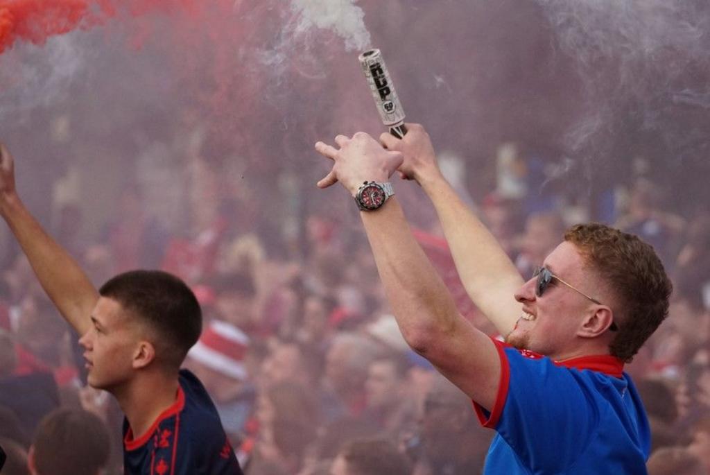 Two young men standing on people's shoulders hold red flares in the air