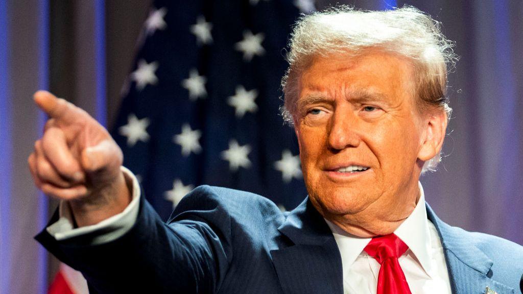 Donald Trump pointing to the right with his right hand raised and pointer finger extended. Shot cuts off just below the shoulder, he's in pin-striped dark suit, white shirt and red tie