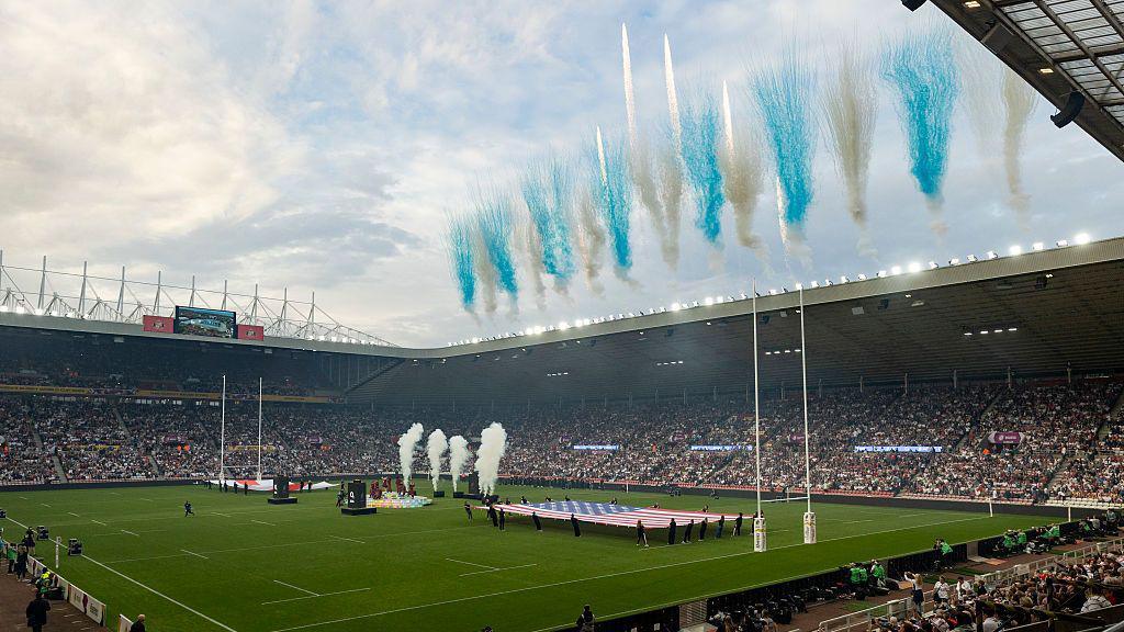 The opening fireworks at the Stadium of Light ahead of the Women's Rugby World Cup 2025. Blue and white fireworks have been set up above the stands, which are filled with thousands of people. A large England and USA flag have been unfurled across the rugby pitch.