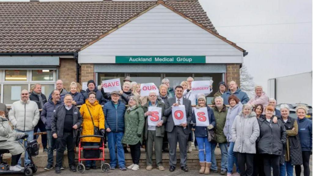 A group of about 30 people are standing outside the Auckland Medical Group GP surgery. It is a single-storey building with large windows and a tiled roof. The people are a mix of men and women in thick jackets, some of whom are holding signs reading "save our surgery" and "SOS".