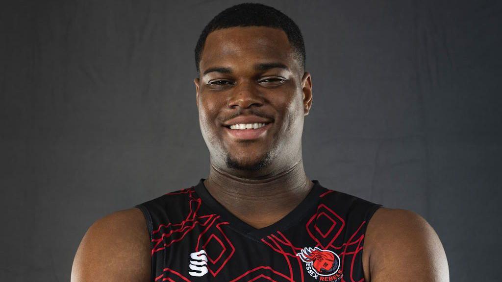 Anthony 'TJ' Hibbert in a black basketball vest with a red pattern and Essex Rebels logo. He smiles at the camera in front of a grey background. 