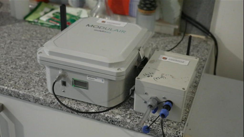 Two grey boxes on a kitchen side. One is larger and reads "modular", it has a black wire coming from it which attaches to a wall behind it. Next to it is a smaller grey box which has a clear tube sticking out of the side, and black wires. They are the machines that have been measuring pollution levels