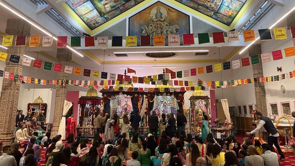 A large crowd of people sit on the carpet inside a temple which is decorated with colourful flags which are hung from the ceiling. The ceiling is painted with Hindu gods.