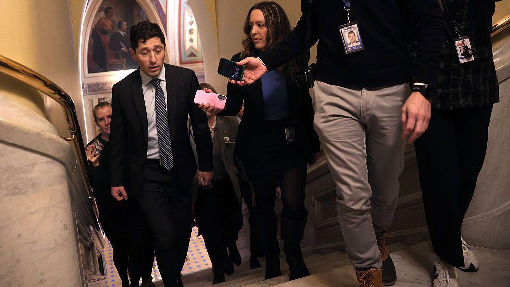 Frey in dark suit and black-and-white tie walks up stairs surrounded by reporters holding their phones out to record him