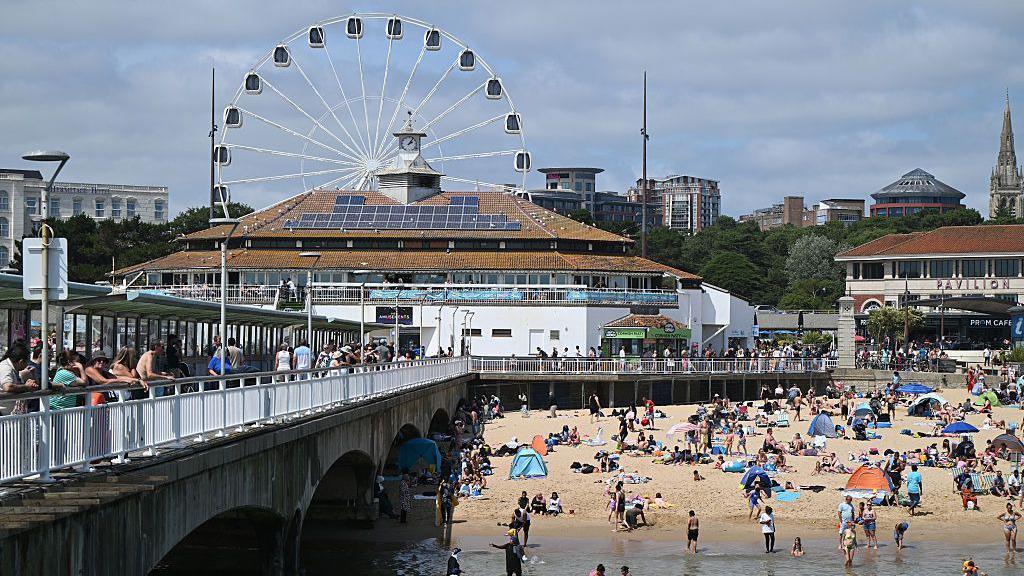 Bournemouth Pier on a sunny day. You can see a big wheel behind a building with solar panels on the roof. There are a lot of people on the beach and on the pier. There is sand and you can see the sea.