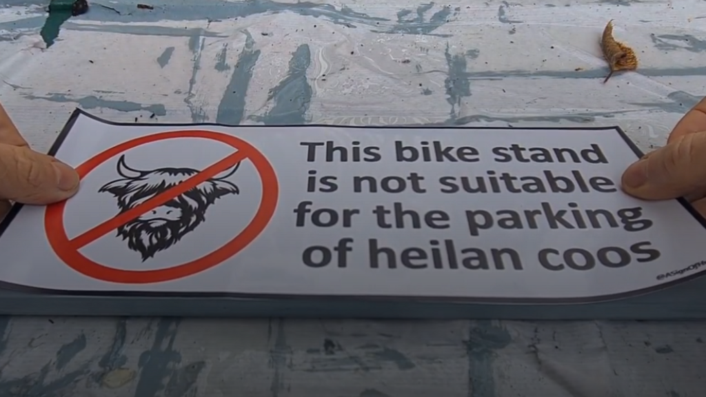 A sign with a circle and cross through a cartoon highland cow head that reads "This bike stand is not suitable for the parking of heilan coos".