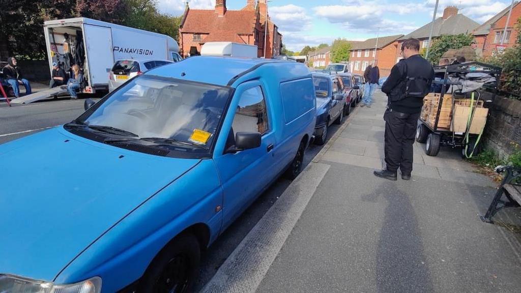 A traffic warden tickets an old blue Fiesta van that is parked on double yellow lines. Across the road is a large film truck with Panavision written across the side. 