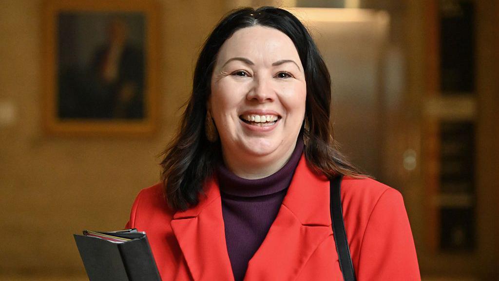 Monica Lennon wearing a bright red blazer over a dark turtleneck top stands indoors holding a black folder. The background is softly blurred, with framed artwork and warm lighting visible.