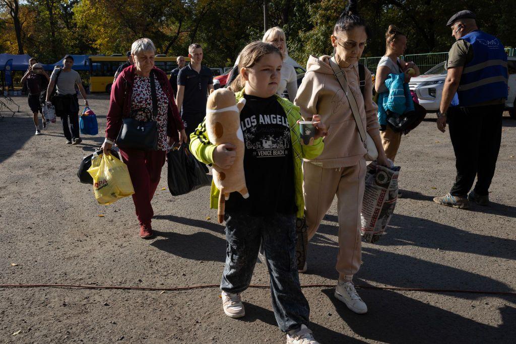 A group of people, including a child or teenager wearinga  yellowy-green jacket, are walking on a gravel car park area, carrying bags.